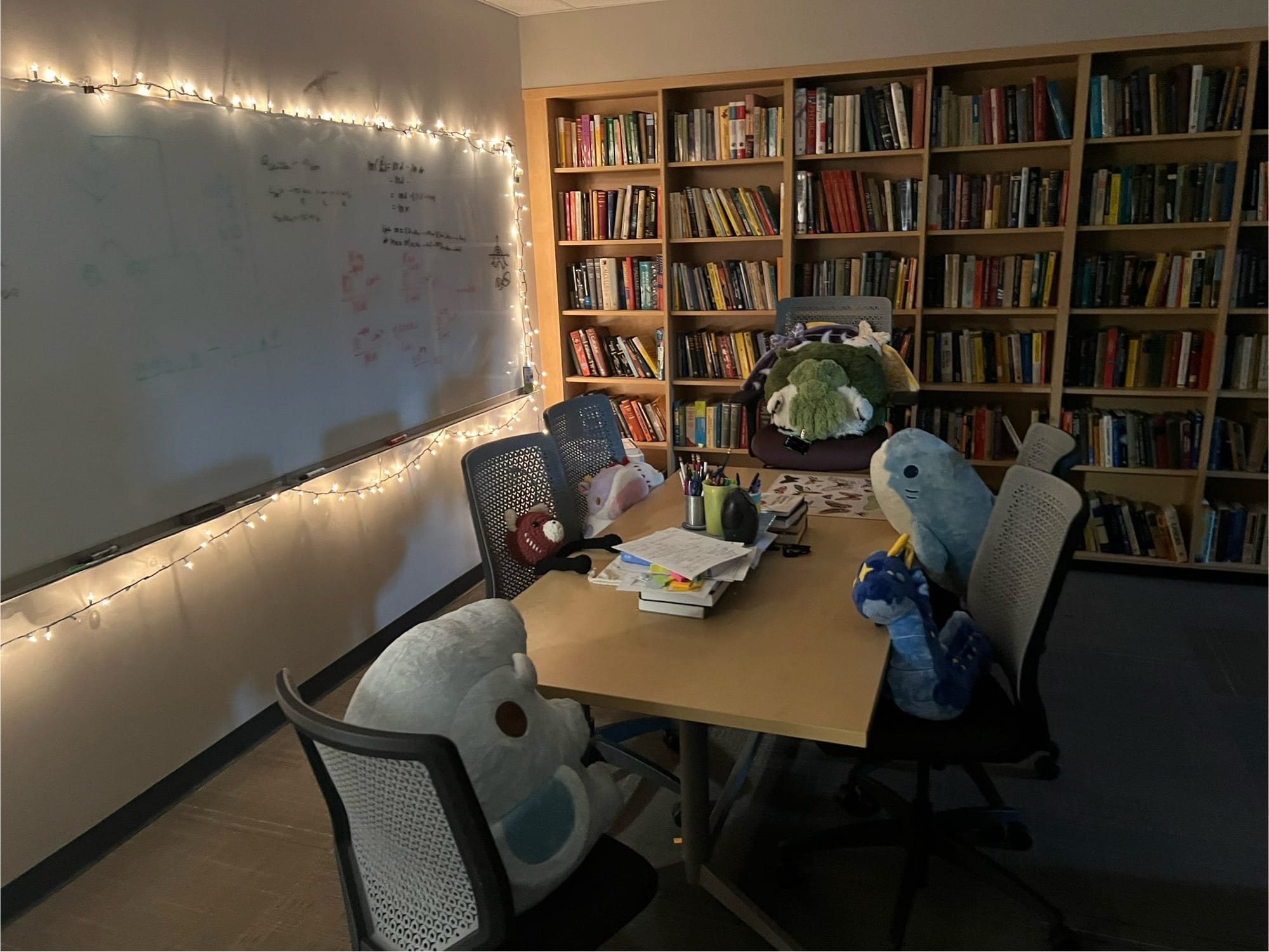 Stuffed animals, including Cthulhu, a shark, a red panda, an elephant, and a dragon sit around a table under string lights surrounding a glassboard with a wall of books in the background.